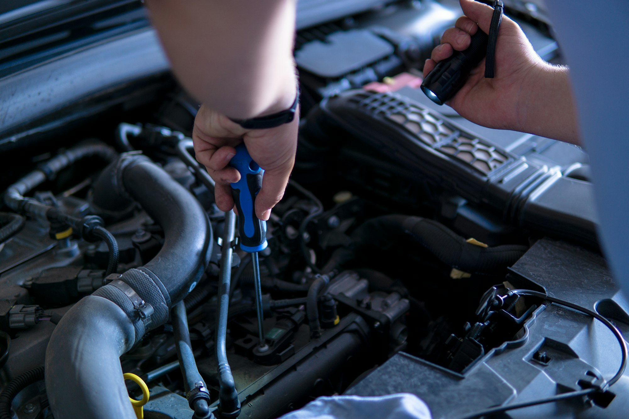 Auto mechanic working and repairing a car engine in a garage with a screwdriver and a torch.